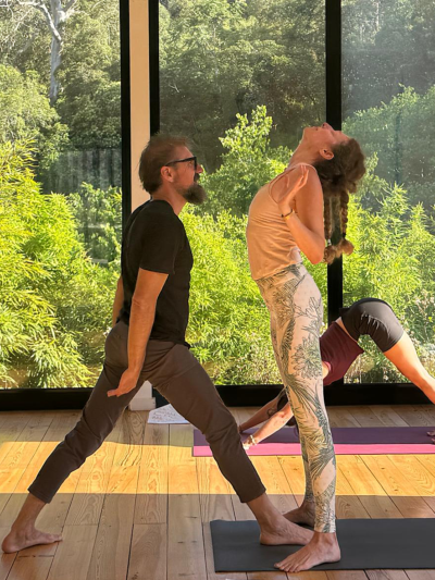 teacher assisting a yoga student to come up from a wheel pose. teacher facing student, student is looking up. forest in the background