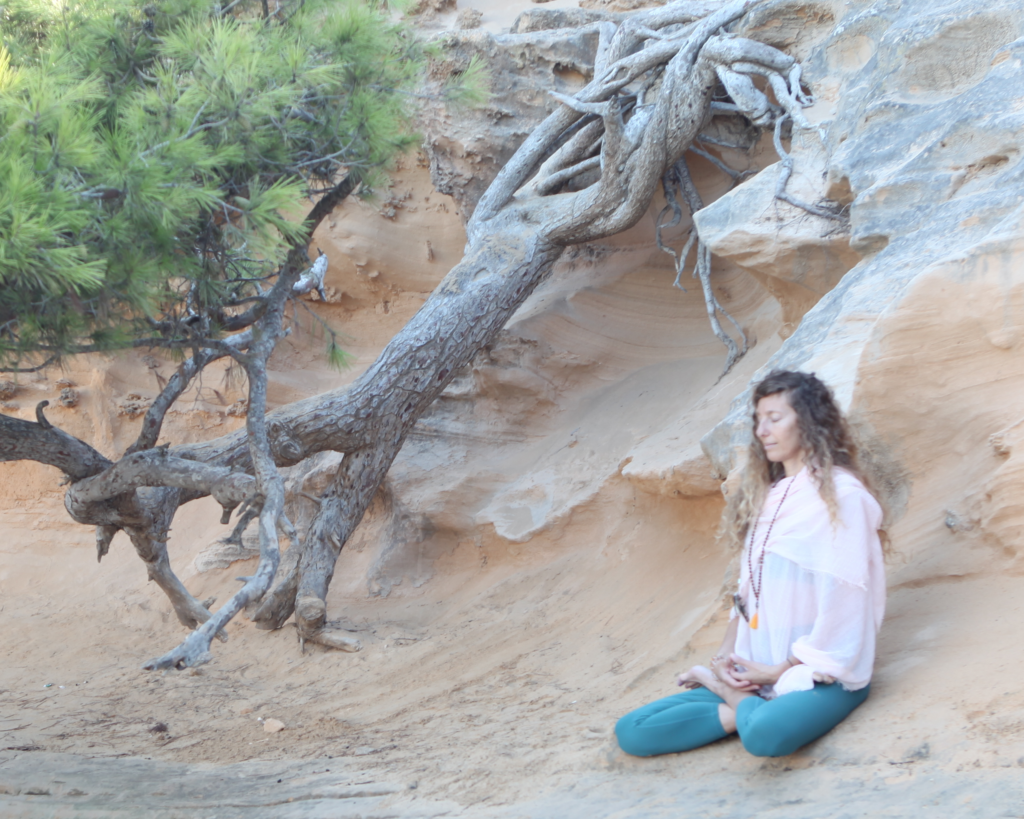 a woman in a seated meditation in nature, against a rocky wall and alongside a pine tree
